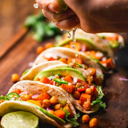 Tacos filled with vegetables and chickpeas on a wooden board, with a hand squeezing lime over them for added flavor.