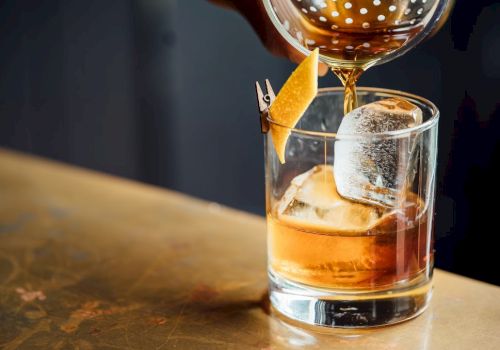 A cocktail being poured over ice in a glass, garnished with a twist of citrus peel clipped to the rim.