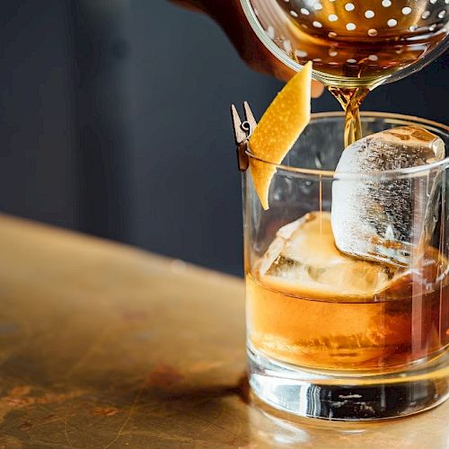 A cocktail being poured over ice in a glass, garnished with a twist of citrus peel clipped to the rim.