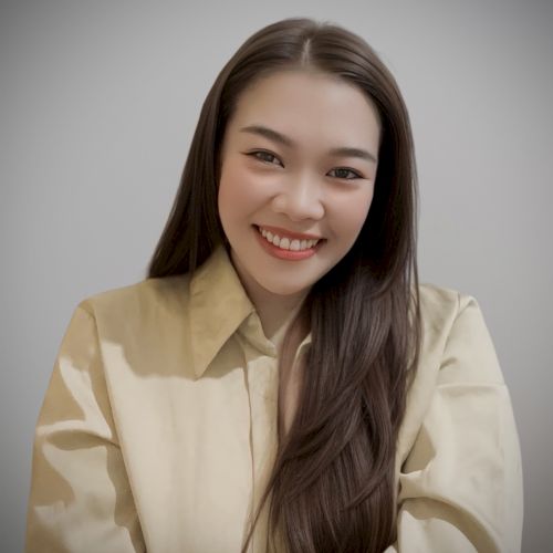 A person with long hair is smiling, wearing a light-colored shirt, against a plain background.