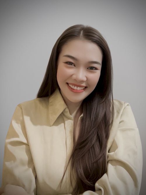 A person with long hair is smiling, wearing a light-colored shirt, against a plain background.