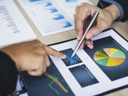 Two people analyzing graphs and charts on a table, pointing with a pen at a colorful pie chart and other data visualizations.