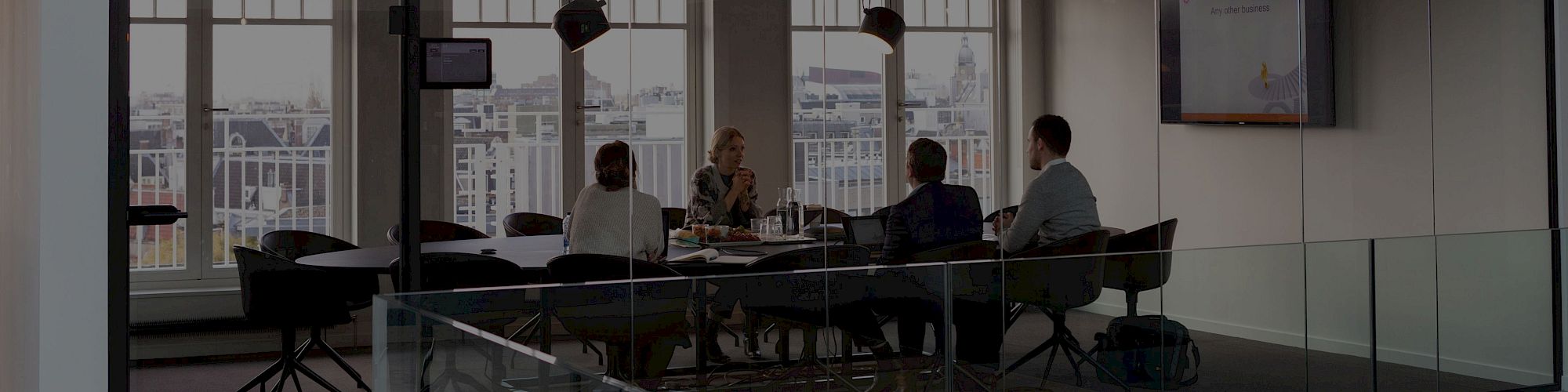 A group of people are in a modern glass-walled conference room having a meeting with a screen displaying content.