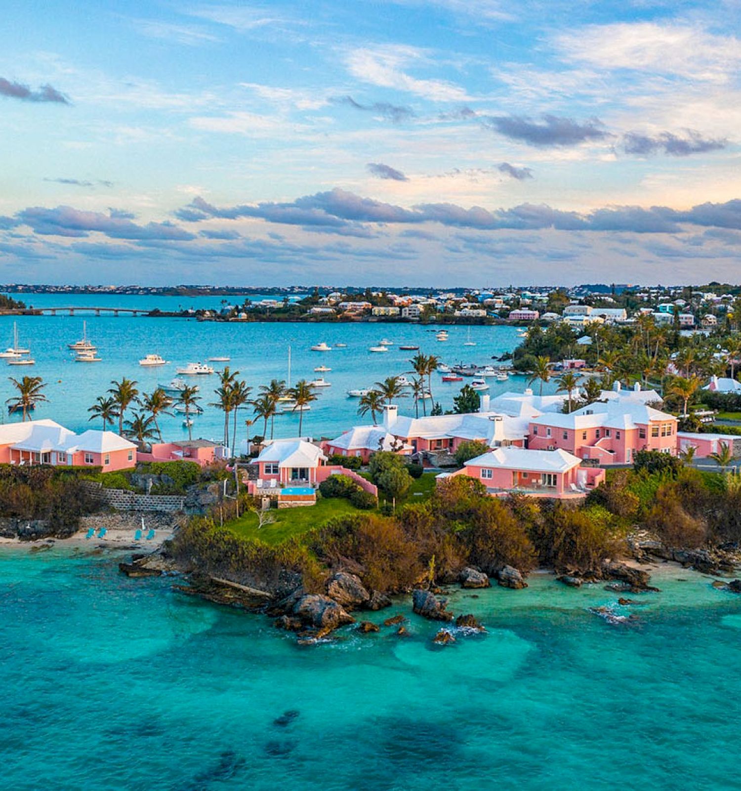 A scenic coastal view featuring turquoise waters, pink houses, lush greenery, and anchored boats under a partly cloudy sky.