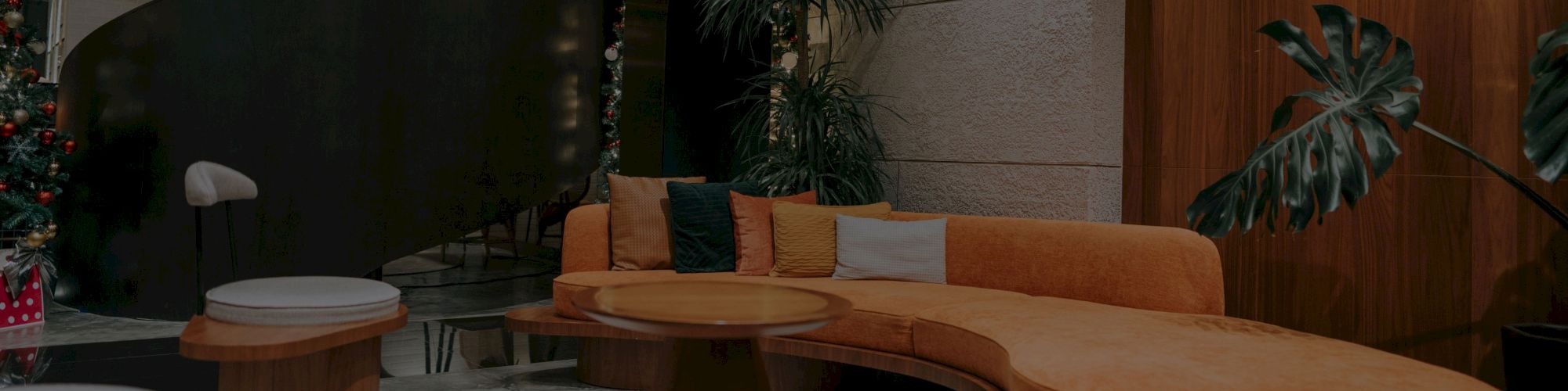 A stylish, modern lounge features a curved sofa, round tables, plants, and subtle lighting, creating a cozy atmosphere.