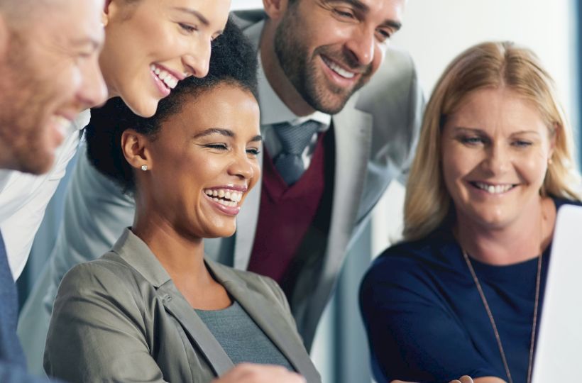 A group of people are gathered around a laptop, smiling and looking engaged, suggesting a positive and collaborative work environment.