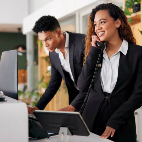 Two individuals in business attire at a reception desk; one talking on the phone, the other focused on a computer screen.
