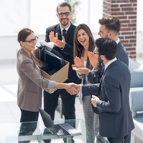 A group of five people in business attire are celebrating, with one person shaking hands and holding a box, symbolizing a new job or promotion.