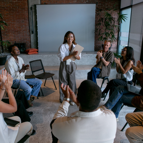 A group of people sit in a circle, clapping for a person standing in the center who is holding papers, in a meeting room setting.