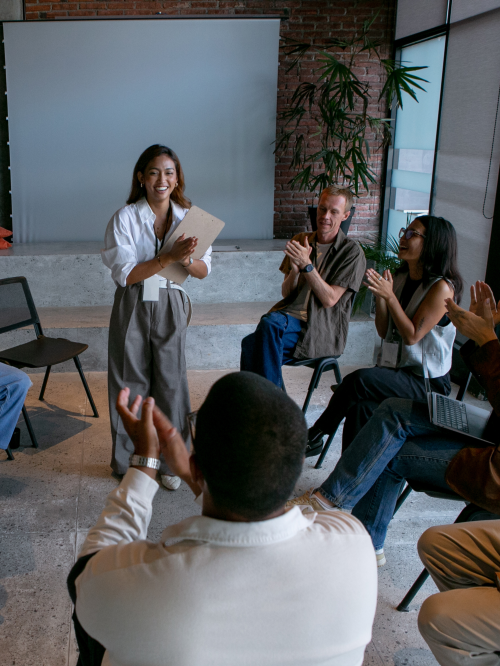 A group of people sit in a circle, clapping for a person standing in the center who is holding papers, in a meeting room setting.