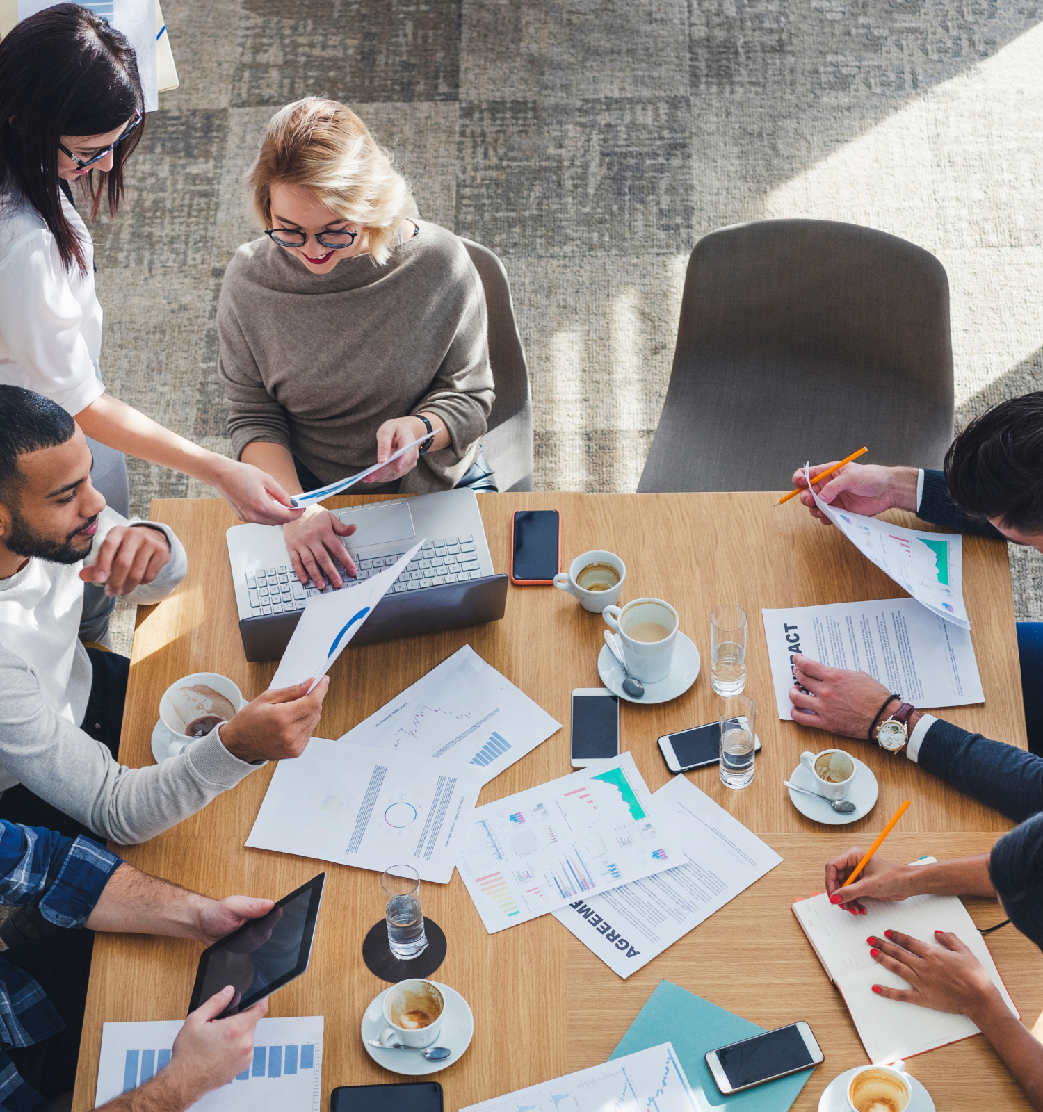 A group of people are gathered around a table with papers, laptops, and coffee, engaged in a collaborative work meeting.