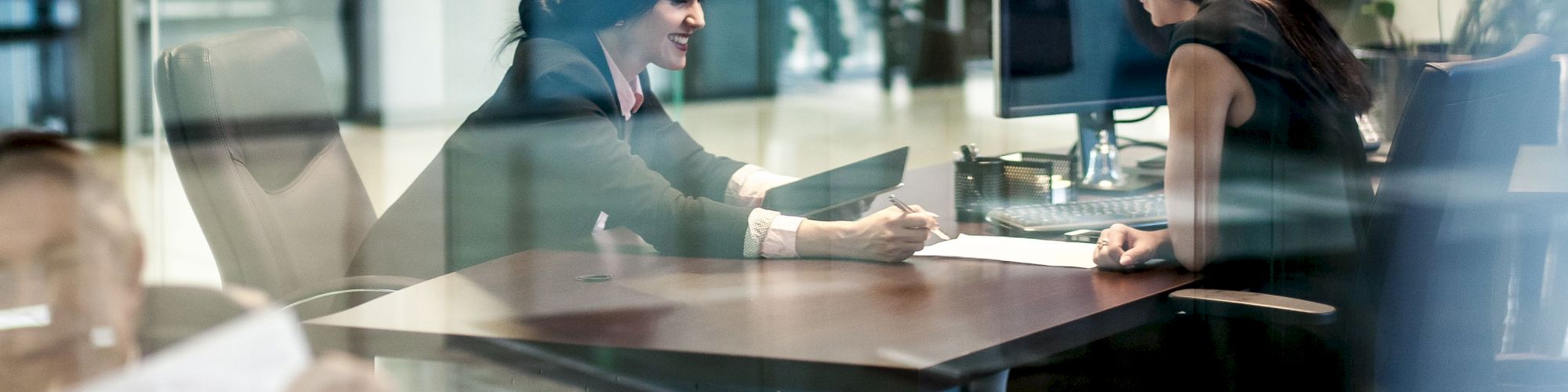 Two people are engaged in a meeting at an office desk. One person is holding a tablet while they both appear focused and engaged.